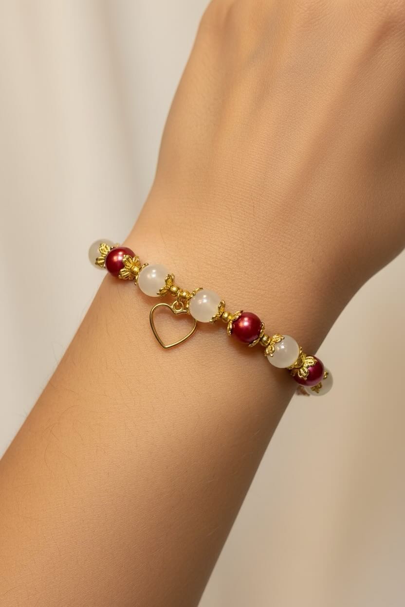A close-up of a gold-tone beaded bracelet on a person's wrist, featuring white and red glass pearls and a small gold heart charm.