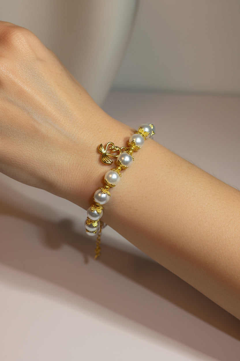 A delicate gold chain bracelet with white glass pearls and a small gold double swan heart charm, shown worn on a person's wrist.