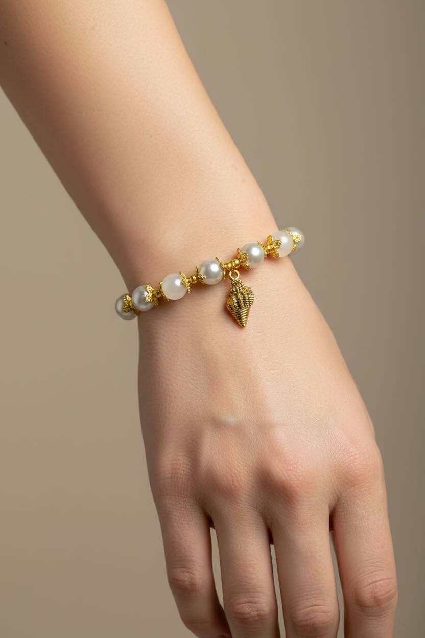 A close-up of a person's wrist wearing a delicate bracelet made of white and silver glass pearls and gold-toned beads with a small gold shell charm, set against a solid warm beige background.
