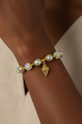A close-up of a gold bracelet with white and silver glass pearls and a small gold conch shell charm, worn on a person's wrist.