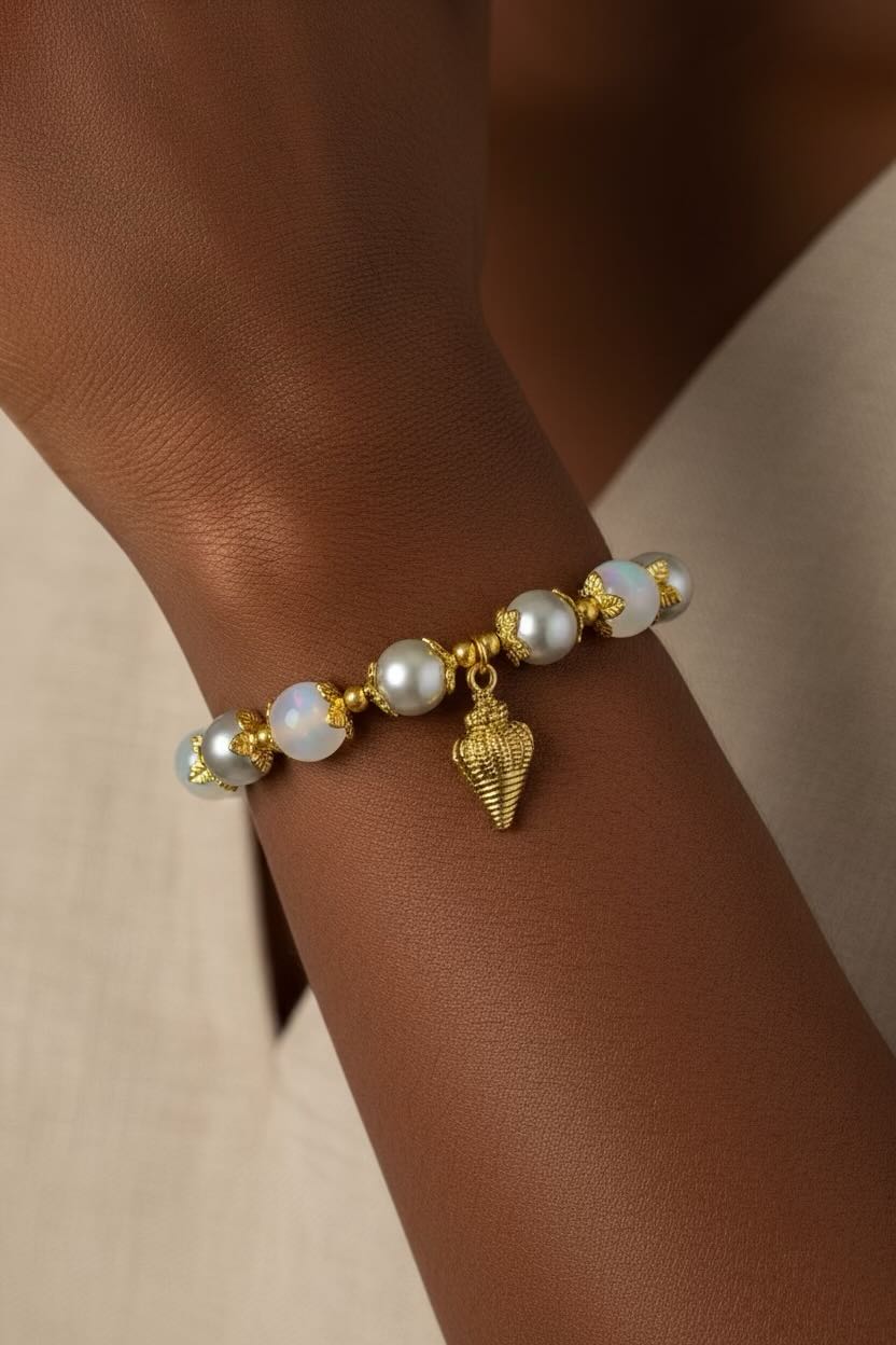 A close-up of a gold bracelet with white and silver glass pearls and a small gold conch shell charm, worn on a person's wrist.
