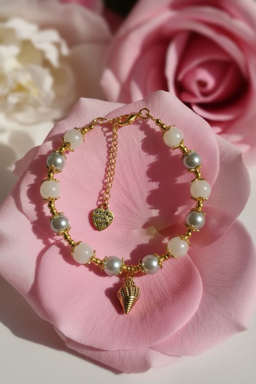 A gold chain bracelet with white and silver glass pearls and a small seashell charm, displayed on a pink rose petal with blurred pink and white roses in the background.