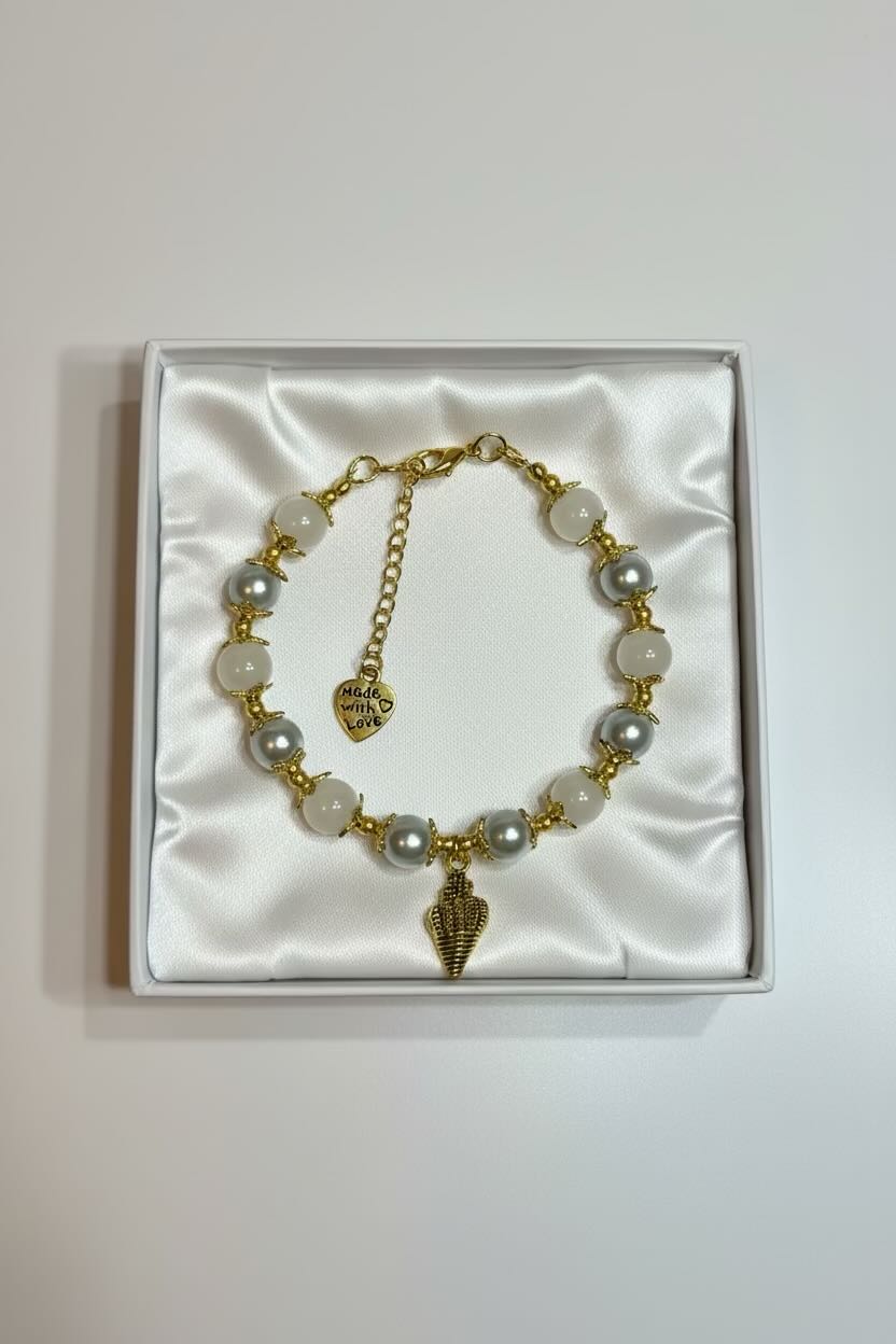 A gold-tone pearl bracelet with white and silver glass pearls and a seashell charm, presented inside a white satin-lined gift box.