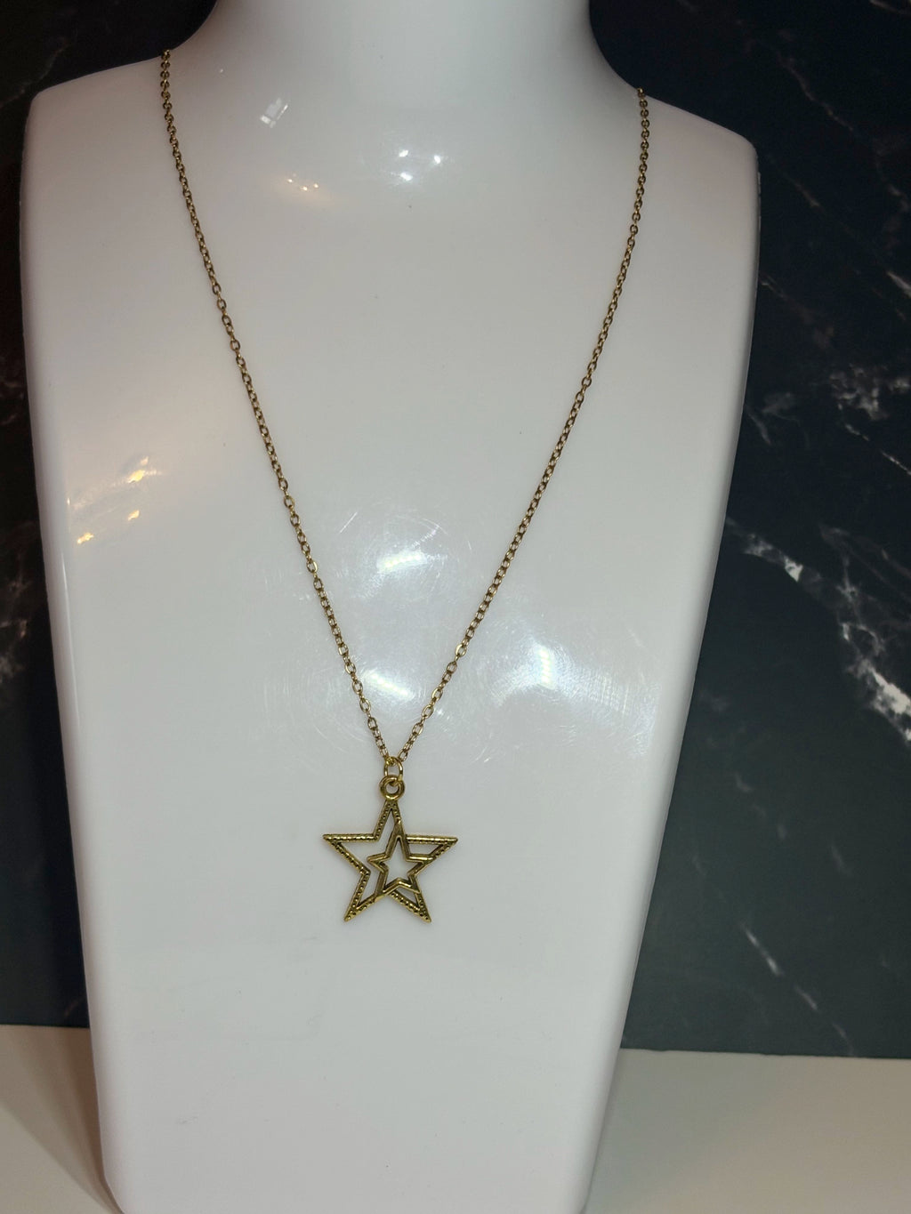 A gold-toned star pendant necklace displayed on a white jewelry bust against a dark marble-patterned background.