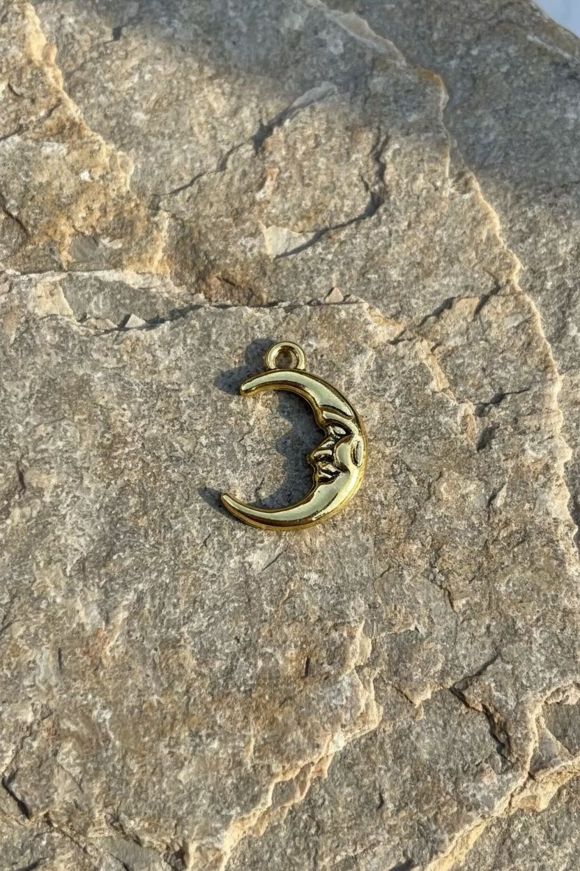 A small crescent moon face charm with a face, finished in antique gold, resting on a textured gray stone surface.
