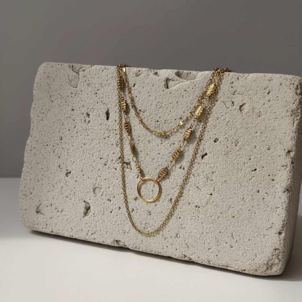 A gold-toned multi-strand necklace with lip chain, leaf style and chain link with a larger circular pendant, displayed on a white mannequin bust against a gray background.