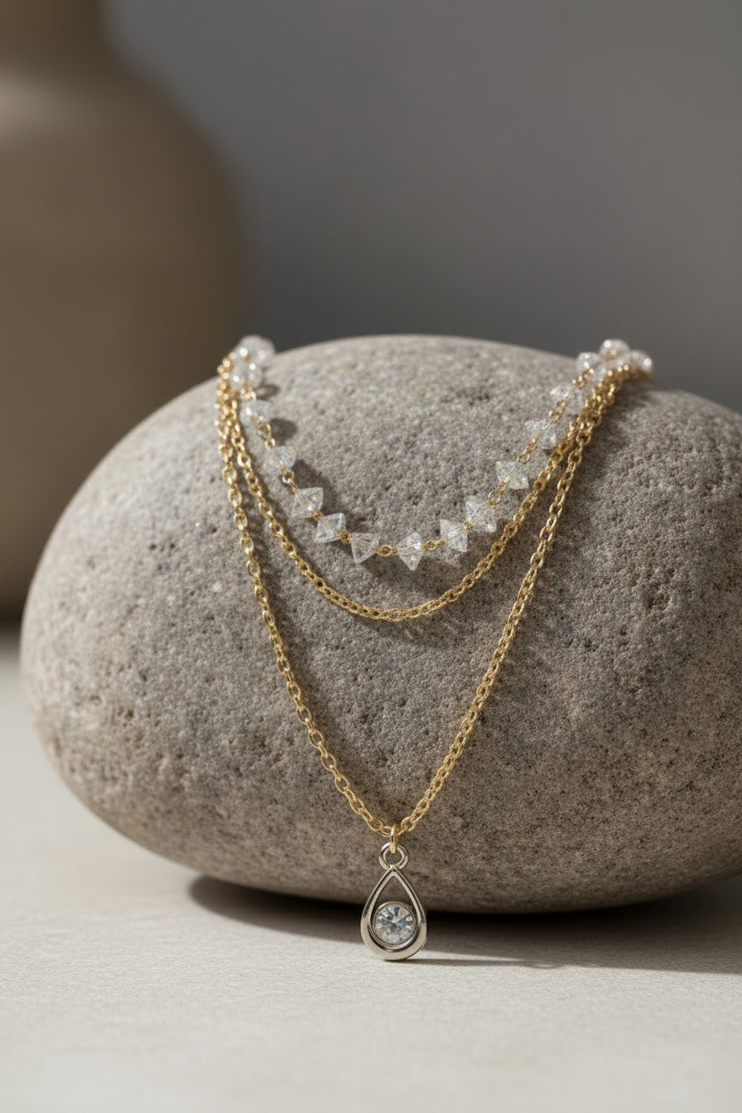 A layered necklace set displayed on a white mannequin bust, featuring a long gold chain with a teardrop pendant set with clear stones, a shorter gold chain, and a delicate clear glass triangle beaded chain.