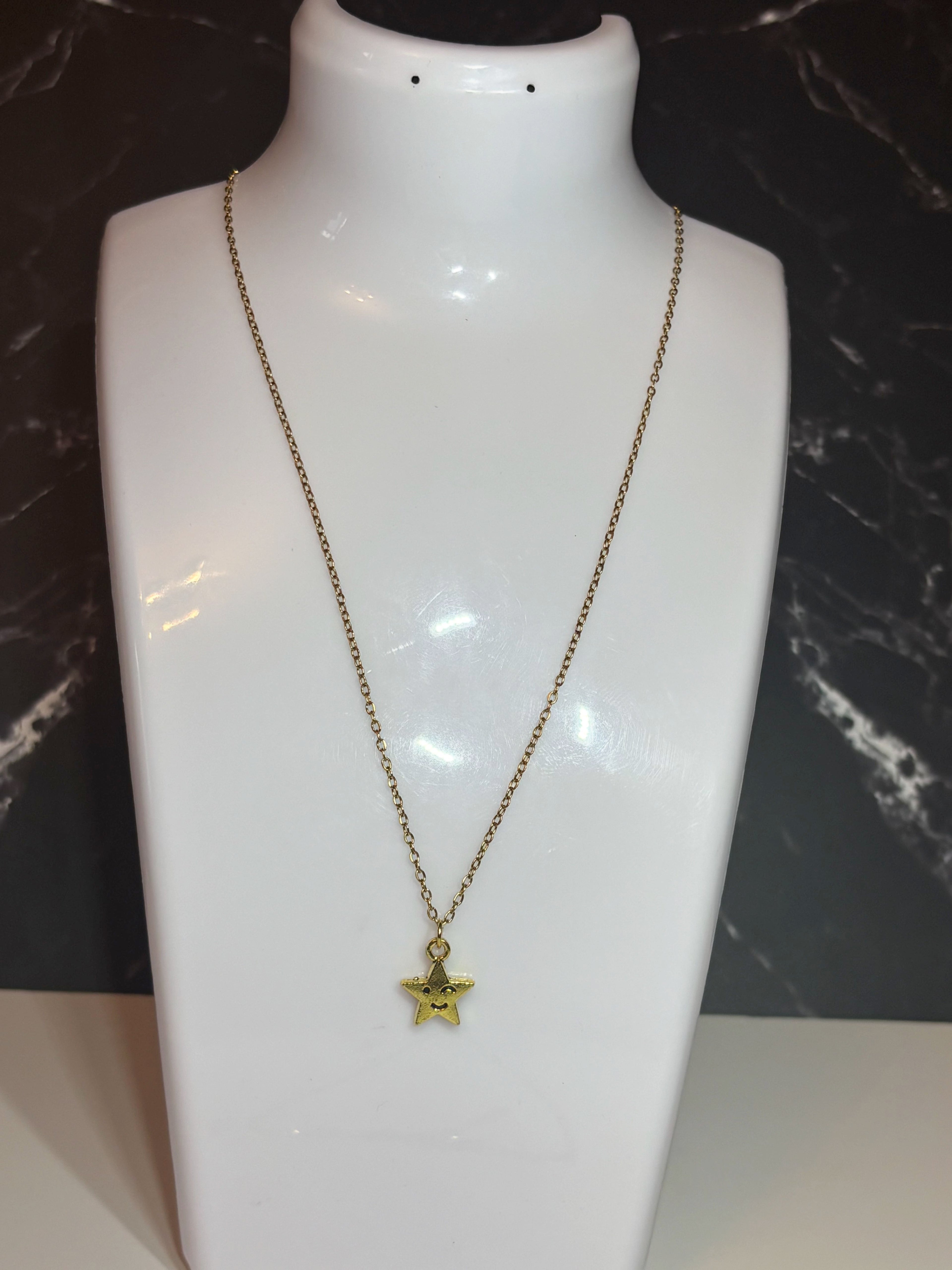 A gold star pendant necklace on a delicate gold chain, displayed on a white mannequin bust against a dark marble-patterned background.