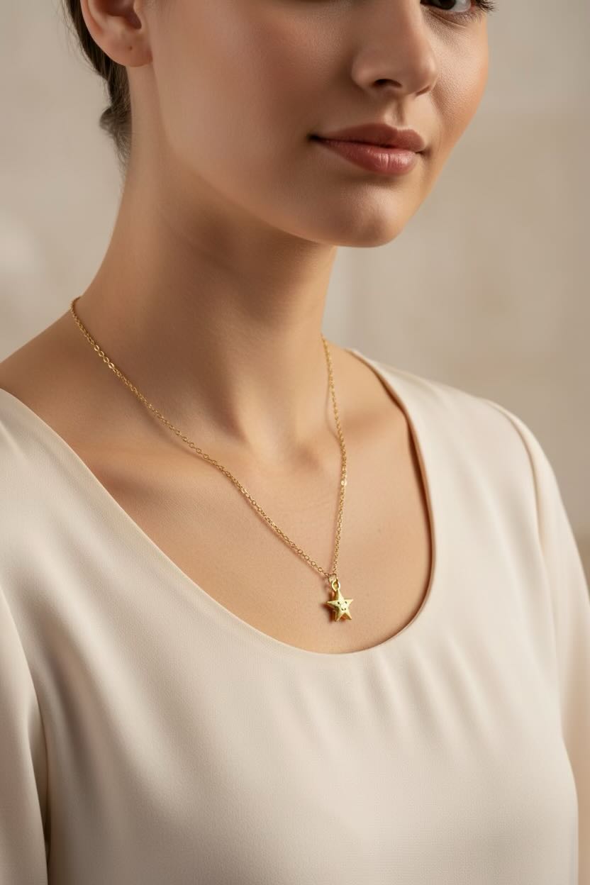 A close-up of a woman wearing a delicate gold chain necklace with a small star pendant, paired with a cream-colored scoop-neck top against a soft beige background.