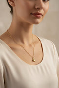 A close-up of a woman wearing a delicate gold chain necklace with a small star pendant, paired with a cream-colored scoop-neck top against a soft beige background.