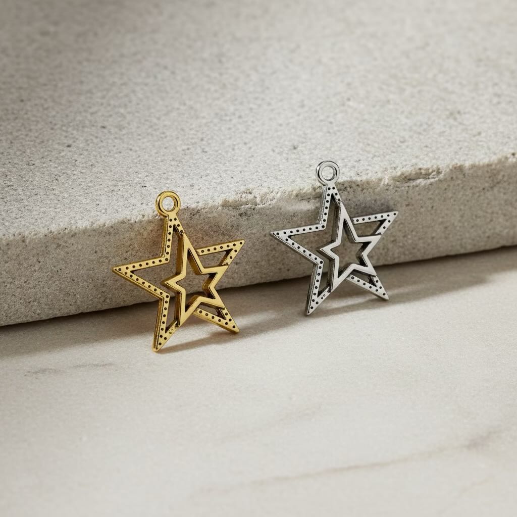 Two small star-shaped jewelry charms, one gold-toned and one silver-toned, displayed on a light beige stone surface.