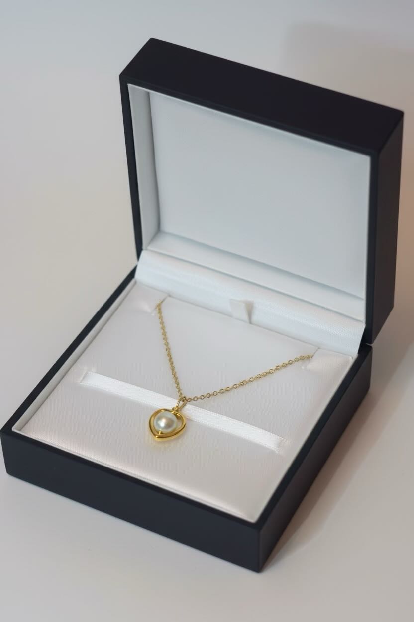 A gold heart pendant necklace with an aqua blue pearl center, displayed inside an open black jewelry box with a white velvet interior.