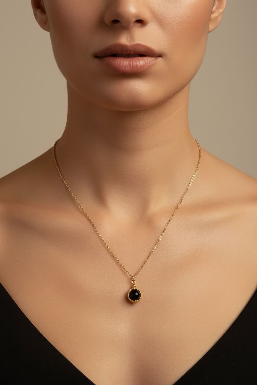 A close-up of a model wearing a delicate gold chain necklace with a small round black pearl pendant, set against a neutral beige background.