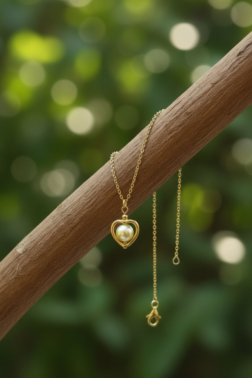 A gold heart-shaped pendant necklace with a white pearl inside, draped over a brown wooden branch against a soft green bokeh background.