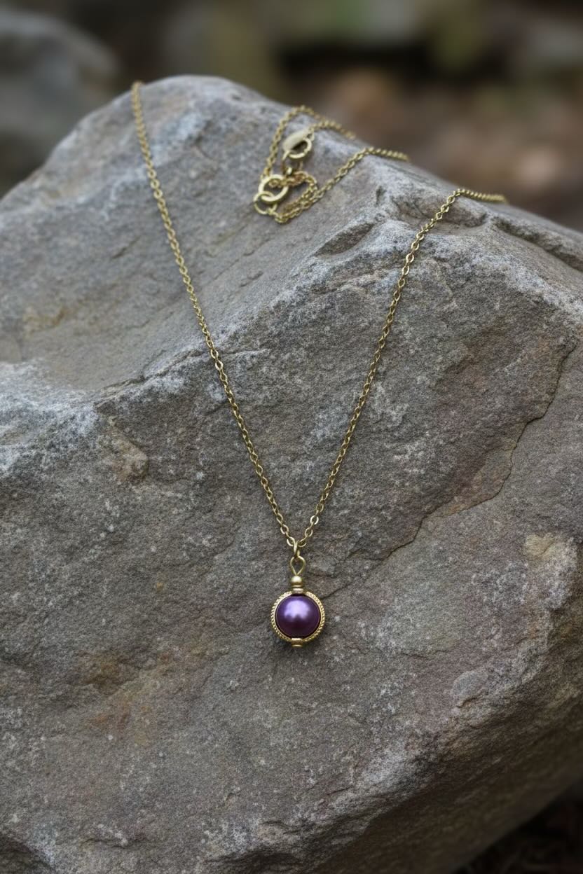 A gold chain necklace with a round dark orchid purple pearl pendant resting on a textured gray rock outdoors.