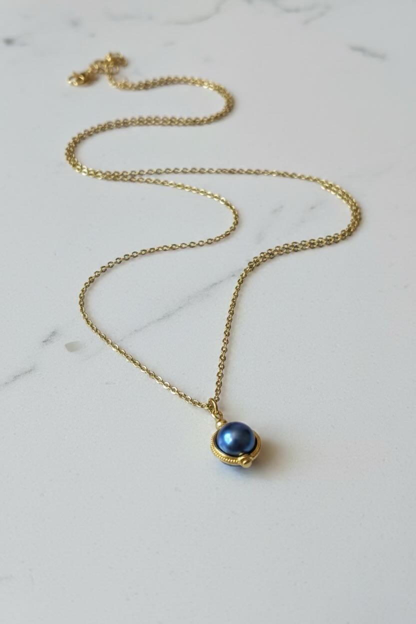 A gold chain necklace with a small oval pendant featuring a dark royal blue pearl set in a gold bezel, displayed on a light marble surface.