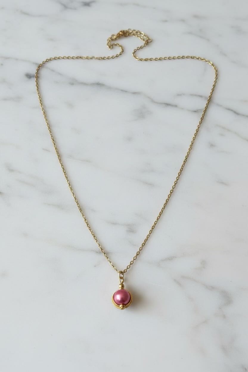A delicate gold chain necklace with a small hot pink pearl pendant in a gold bezel setting, displayed on a white marble surface.