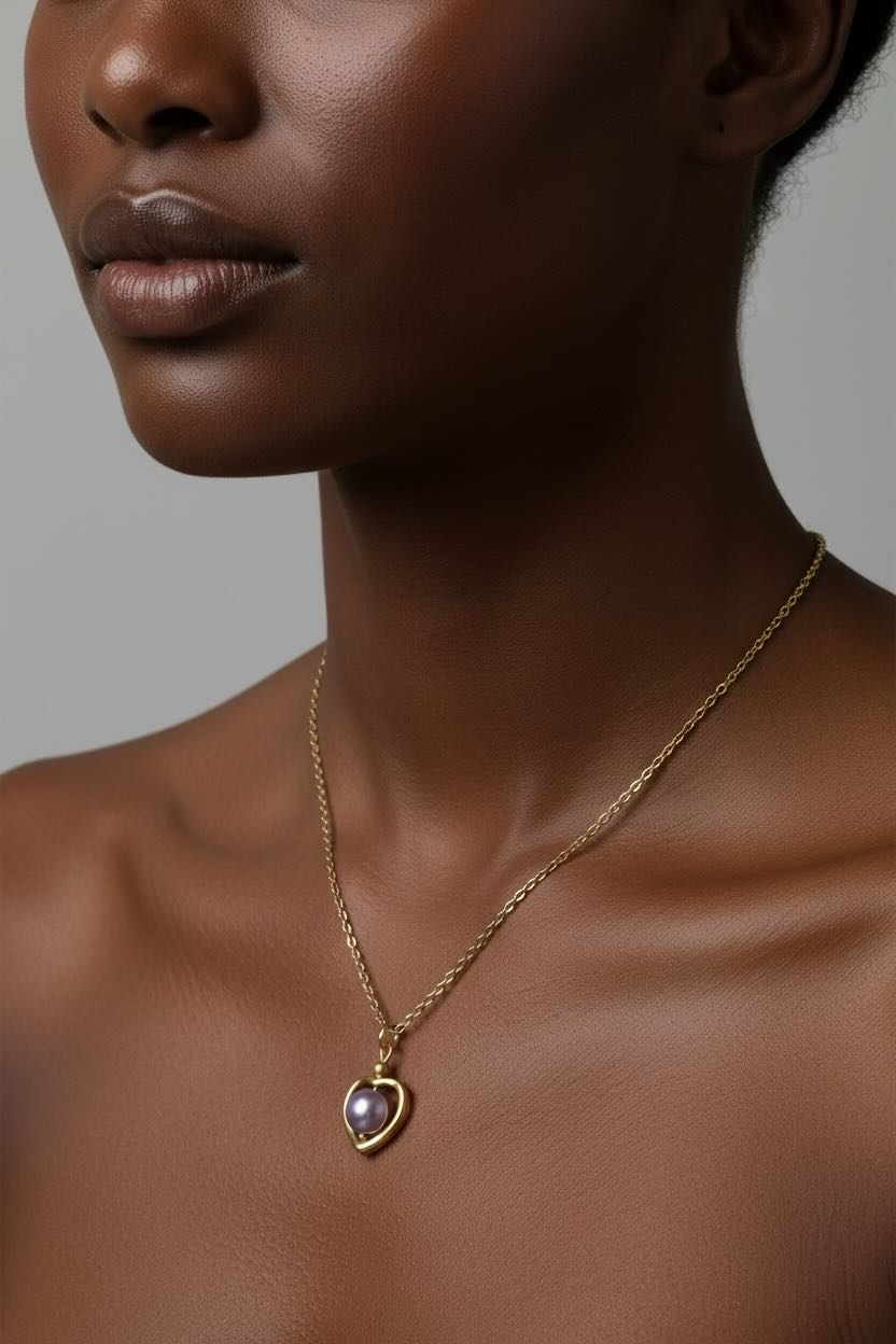 A close-up of a model wearing a delicate gold chain necklace with a heart-shaped pendant featuring a lavender pearl, set against a neutral gray background.