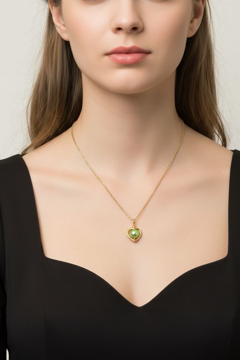 A close-up of a woman wearing a delicate gold chain necklace with a small heart-shaped pendant featuring a light sea green, styled with a black top against a light gray background.