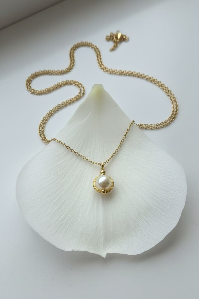 A delicate gold chain necklace with a small gold ring and a pale cream pearl pendant, displayed on a white flower petal with its matching clasp shown above.