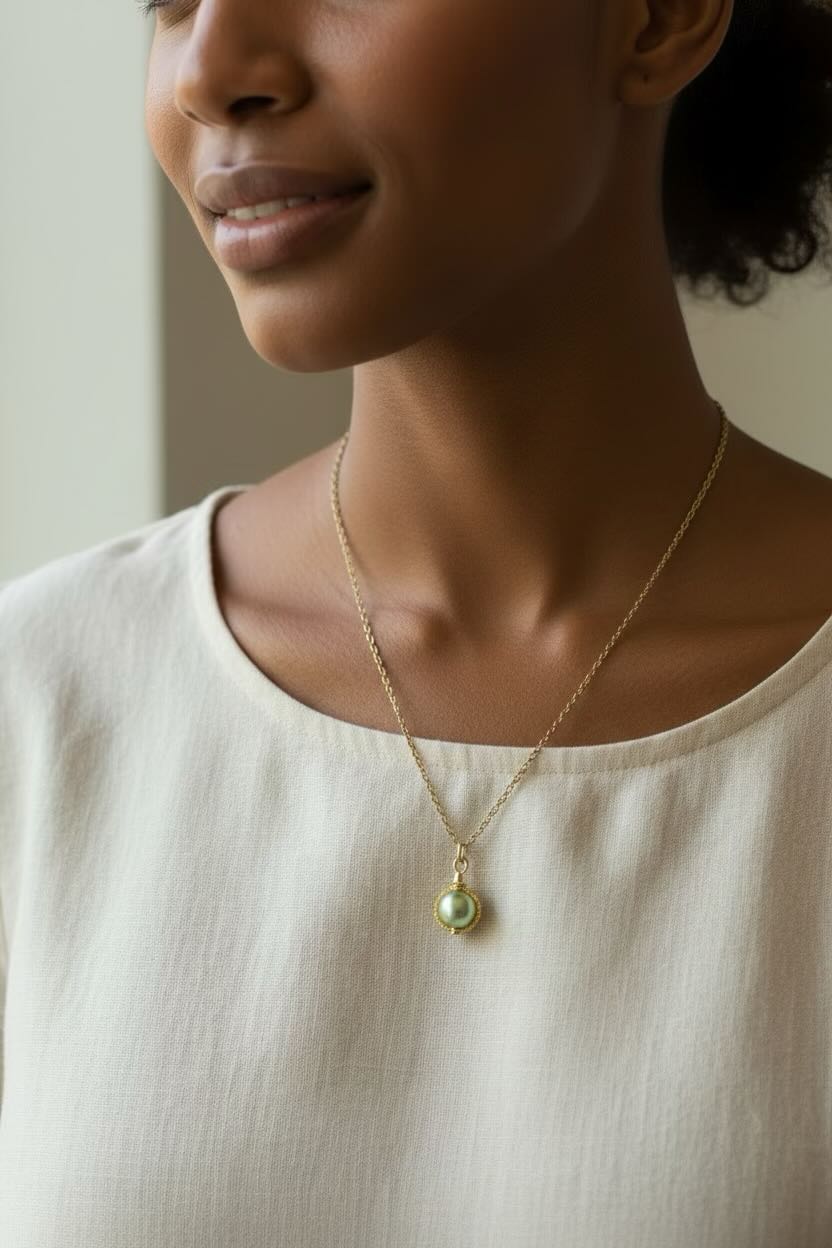 A close-up lifestyle shot of a woman wearing a delicate gold chain necklace with a small, round, sage green pearl pendant, paired with a cream-colored linen top.