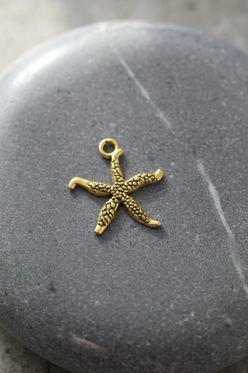 A small gold starfish-shaped jewelry charm with a textured surface and a top loop, resting on a smooth dark gray stone.
