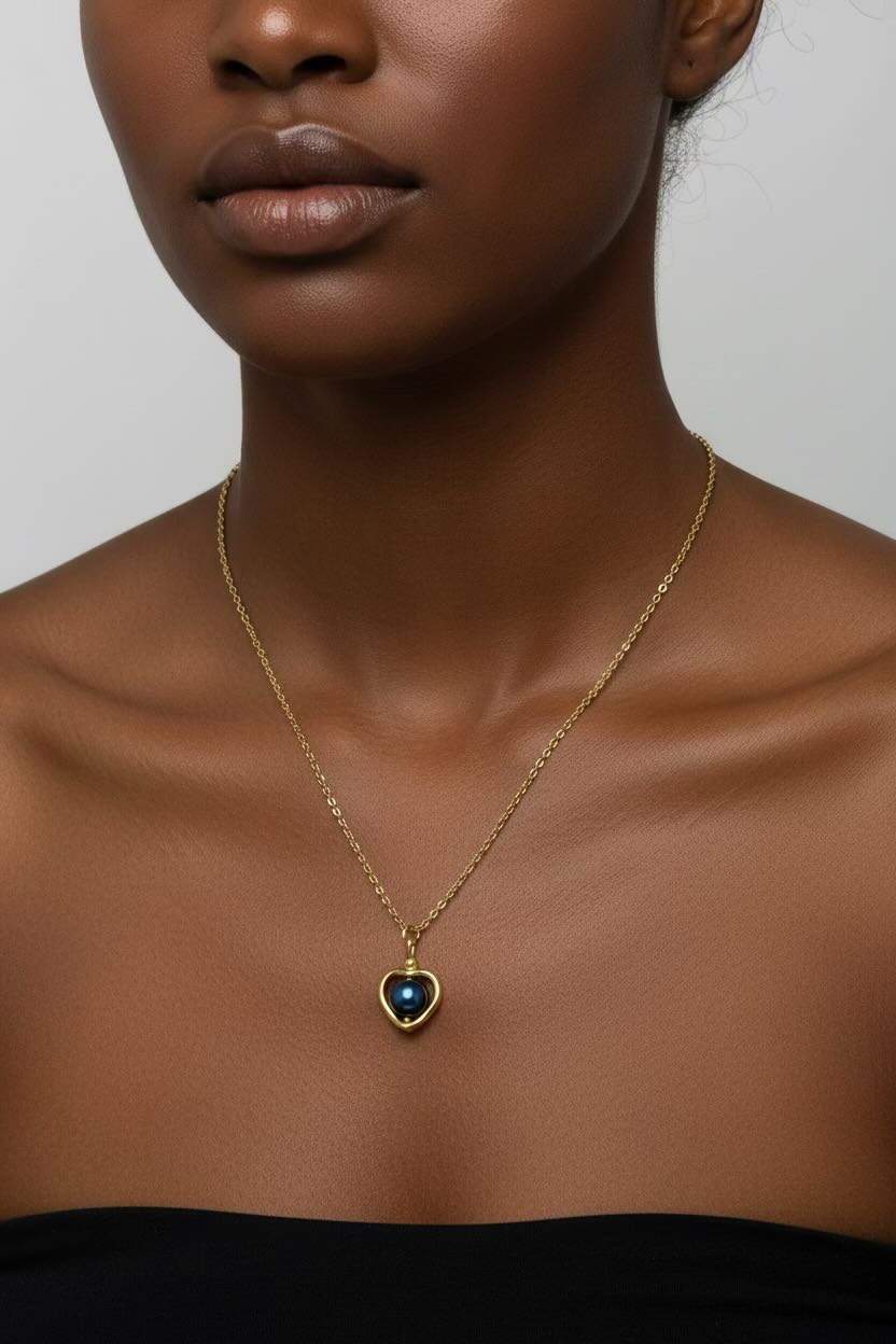 A close-up of a model wearing a delicate gold chain necklace with a heart-shaped pendant featuring a steel blue pearl.