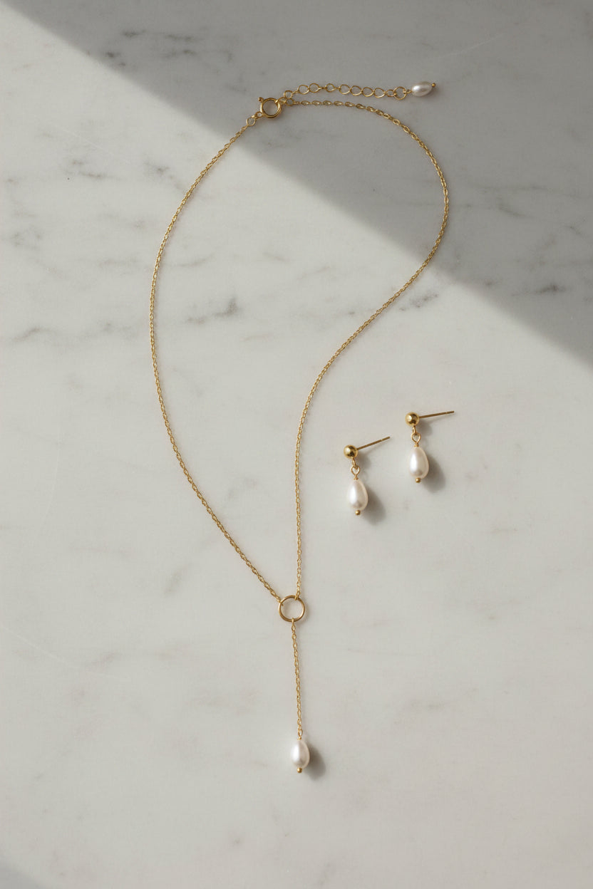 A close-up of a delicate gold Y-necklace with a single pearl drop pendant, paired with a matching pearl stud earring set on a large white marbled surface
