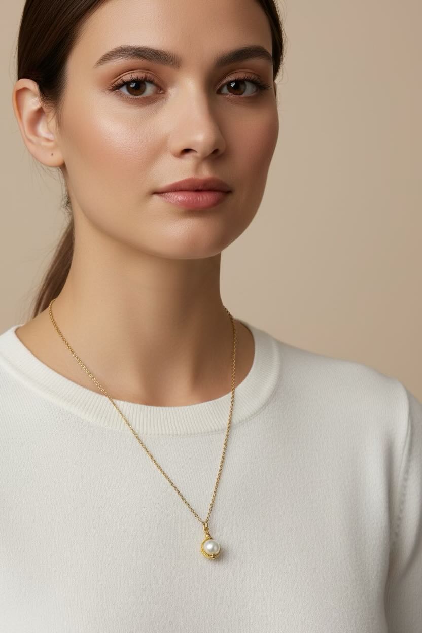 A close-up of a woman wearing a delicate gold chain necklace with a small white pearl pendant, paired with a white crewneck sweater against a soft beige background.
