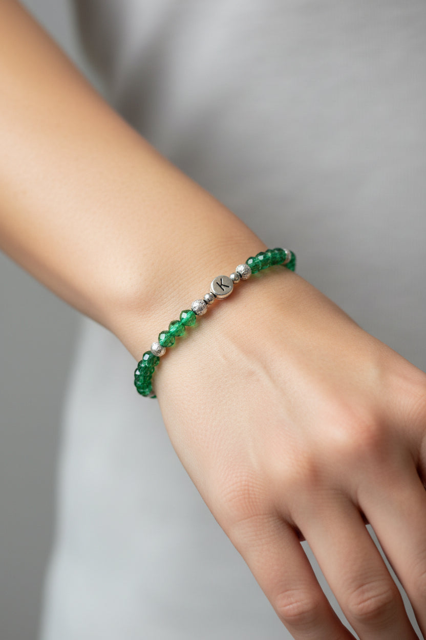 A beaded bracelet with green faceted beads, silver spacer beads, and a central silver 'K'charm engraved with a monogram letter 'K', displayed on a model's arm.