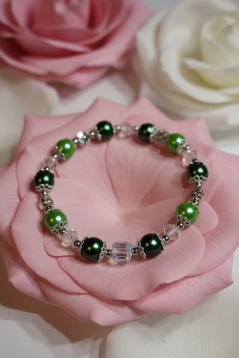 A beaded stretch bracelet with green and dark green glass pearl beads, silver-toned spacer beads, and a clear faceted crystal charm, displayed on a pink rose.