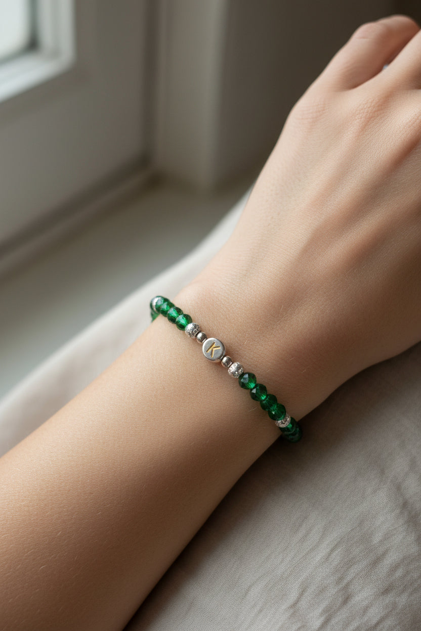 A beaded bracelet with dark green faceted beads, small silver spacer beads, and a central gold-toned bead featuring the letter 'K', displayed on a model's arm.