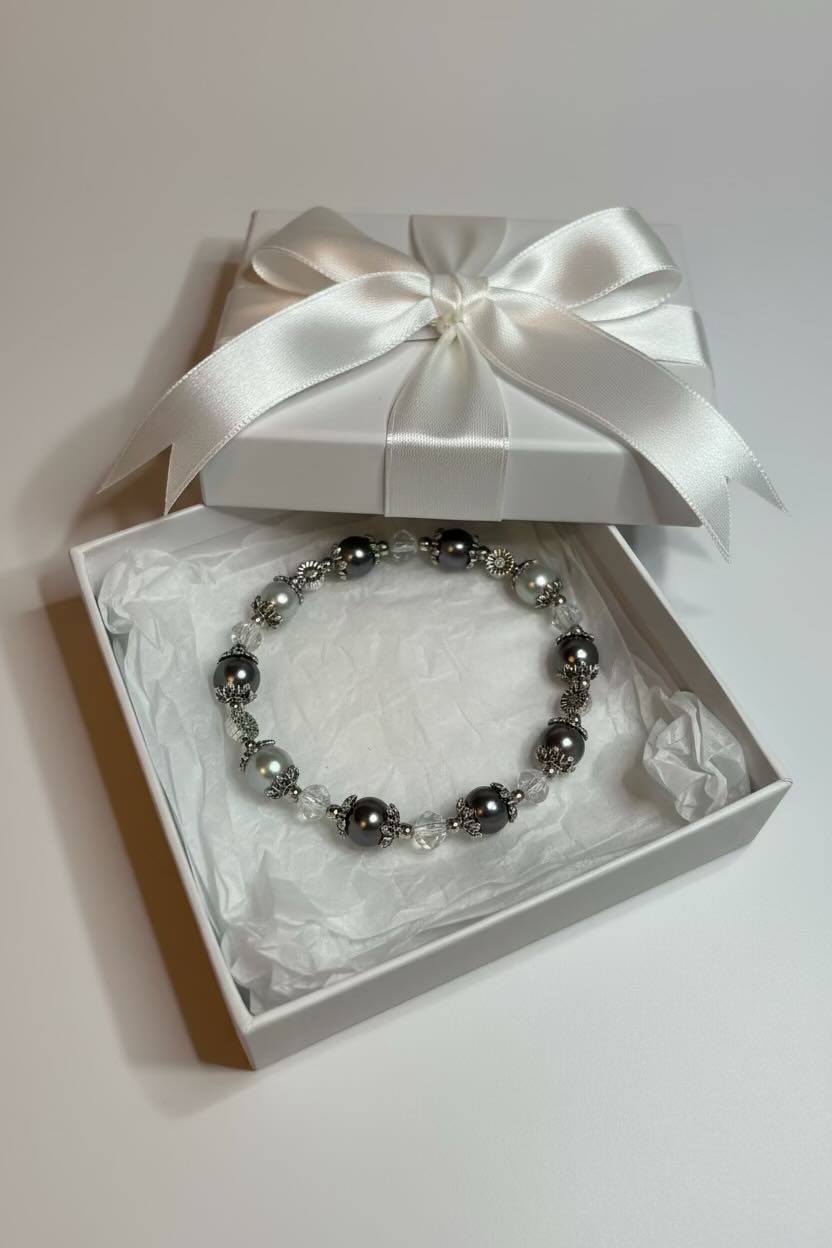 A beaded bracelet with dark gray and silver glass pearls, presented inside an open white gift box with a white satin ribbon bow on a light gray background.