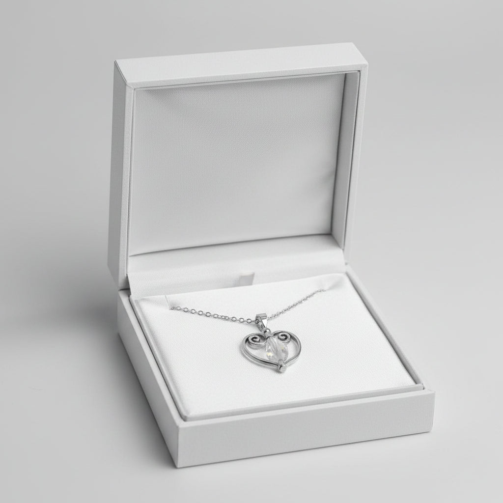 A silver chain necklace with a heart-shaped pendant featuring scrollwork on the left side and a clear faceted bead at the center, displayed in a white gift box.