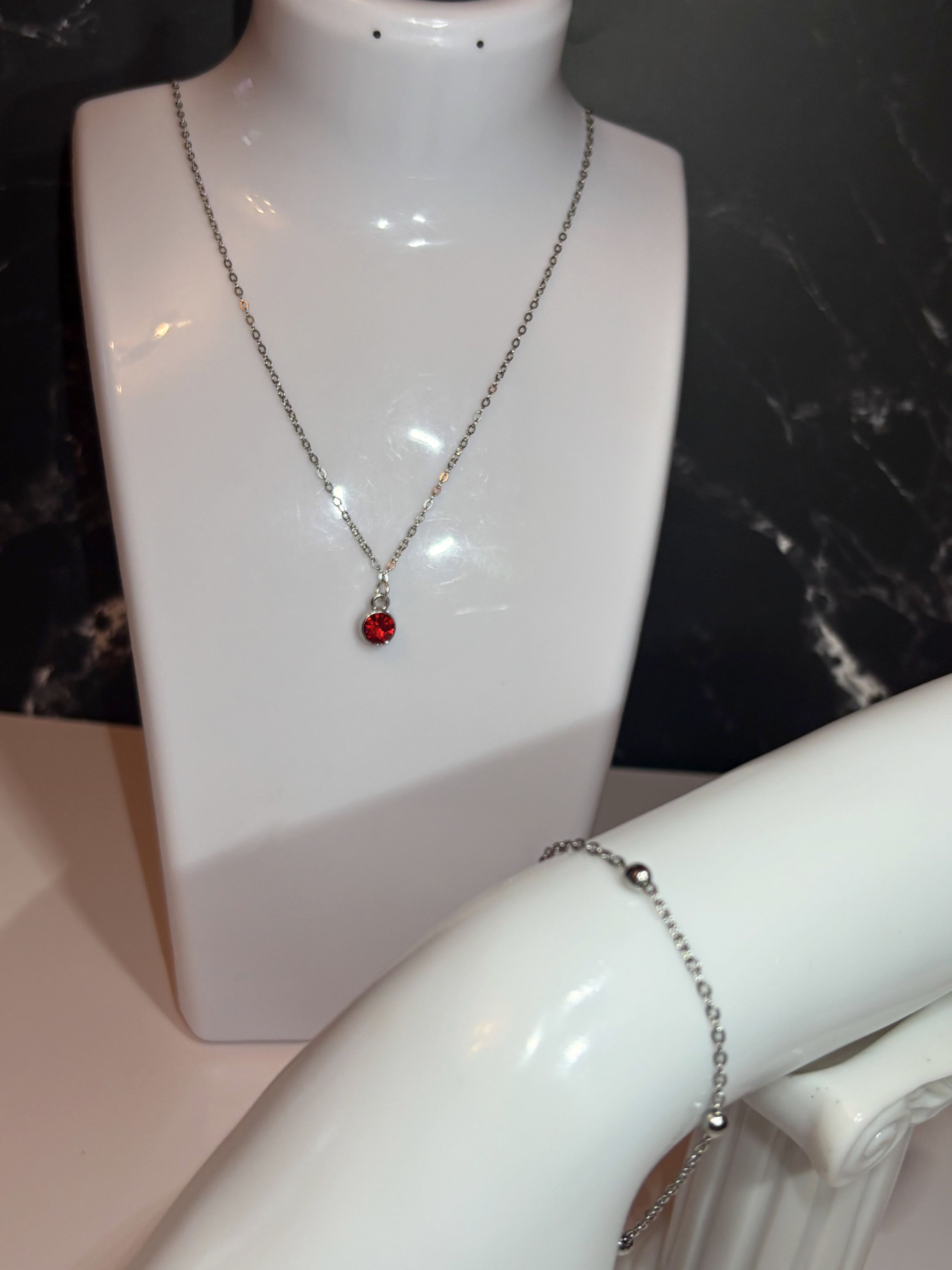 A silver necklace with a small red birthstone ruby pendant is displayed on a white mannequin bust, with a silver ball chain bracelet also visible on a white arm form against a dark marble background.