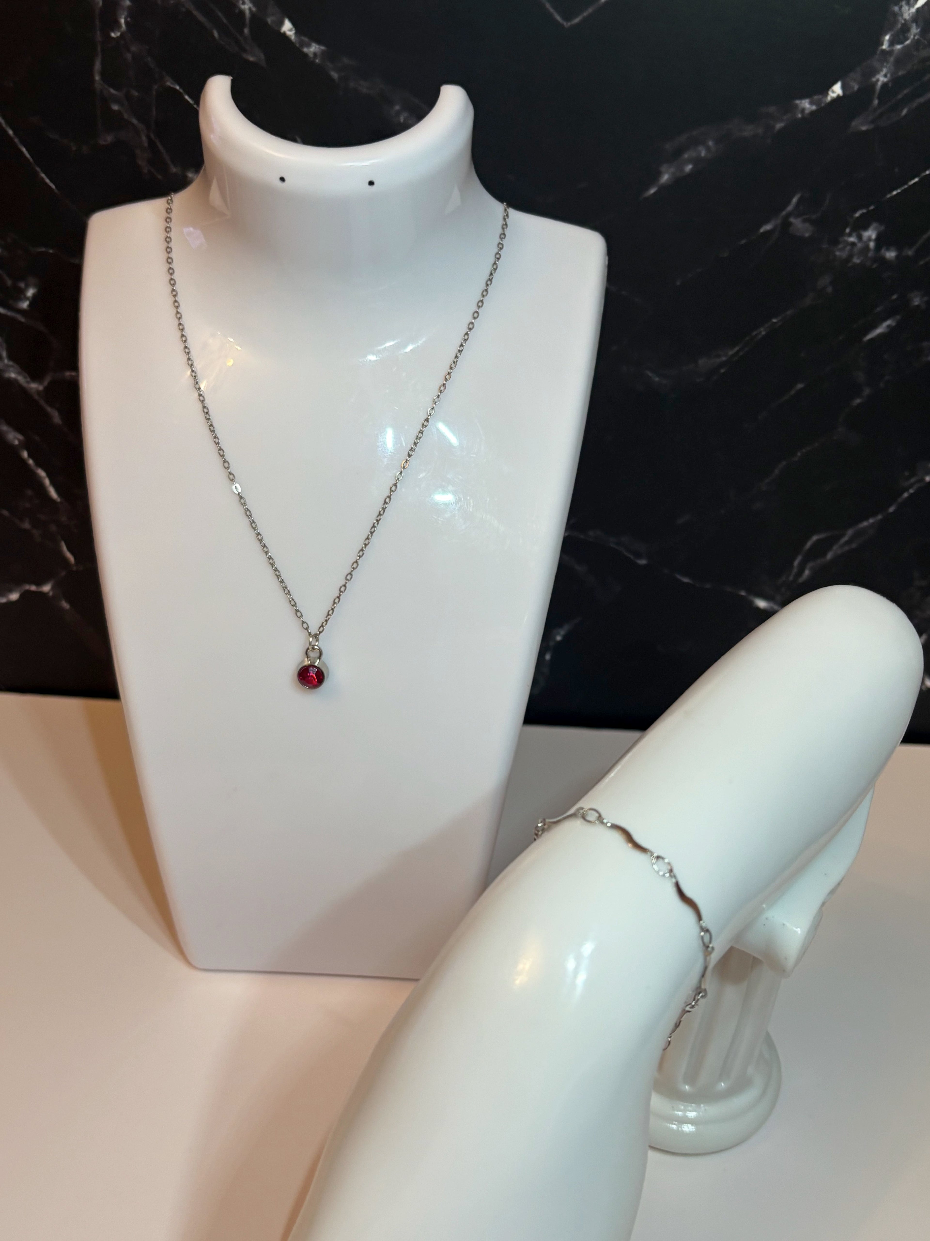 A silver chain necklace with a small red birthstone pendant displayed on a white jewelry bust, alongside a silver wave lined bracelet, set against a black marble background.