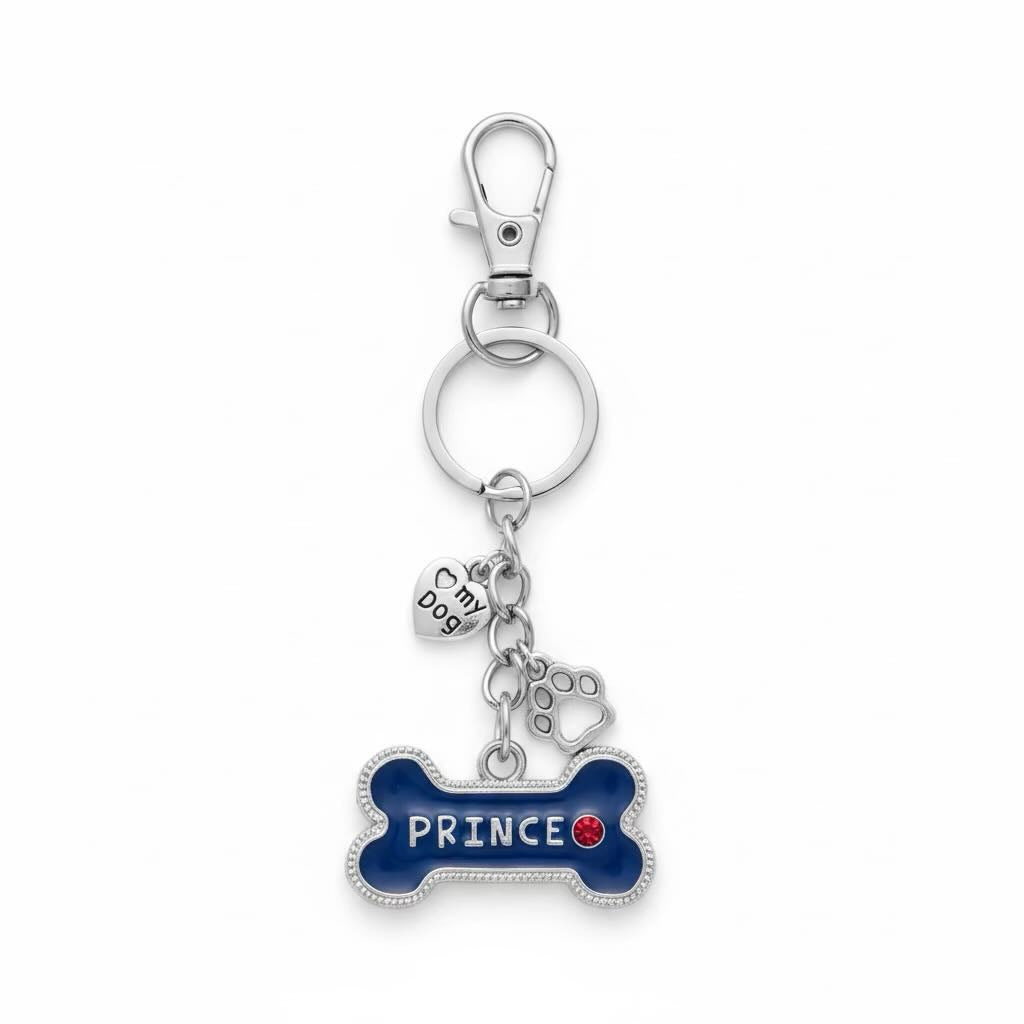 A silver metal keyring/keyrings featuring a bone-shaped charm with blue enamel and the name 'PRINCE' in white, accented with a small red heart. It includes a small paw print charm, a 'My dog' heart charm, and a lobster clasp on a white background.