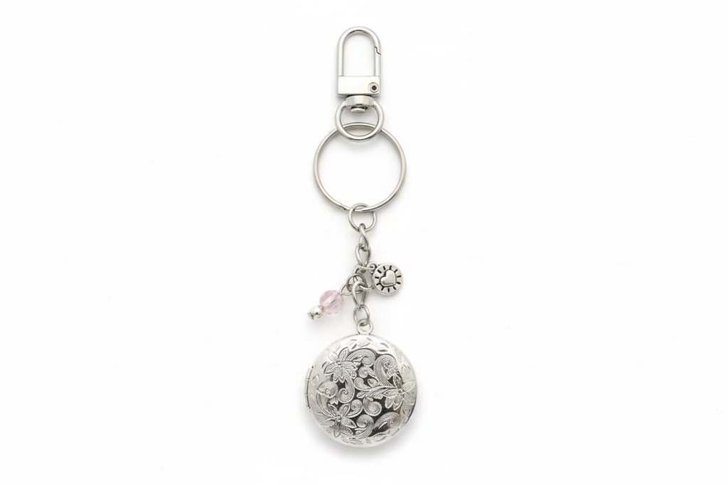 A silver-toned metal keyring featuring a round floral locket pendant, a small pink bead charm, and a silver flower charm, attached to a split ring and lobster clasp on a white background.