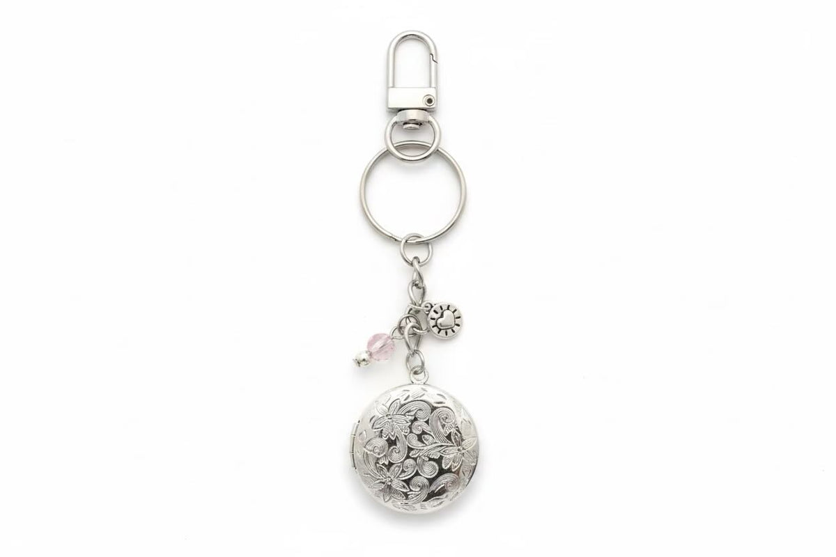 A silver-toned metal keyring featuring a round floral locket pendant, a small pink bead charm, and a silver flower charm, attached to a split ring and lobster clasp on a white background.