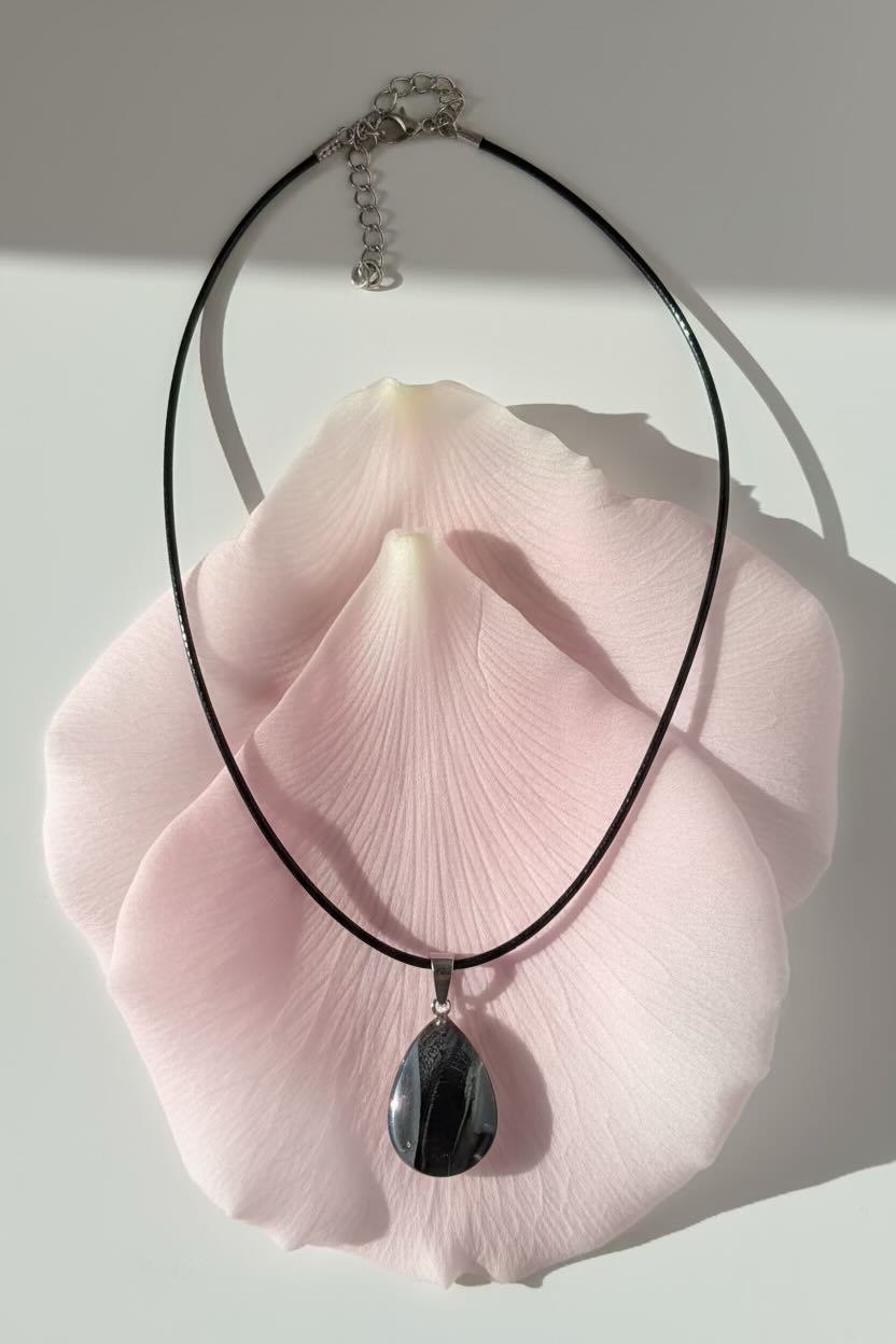 A black cord necklace with a dark black and clear teardrop pendant, displayed draped over a large pale pink flower petal on a white background.