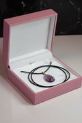 A purple teardrop-shaped amethyst pendant necklace on a black cord, presented inside an open pink jewelry box with a white interior.