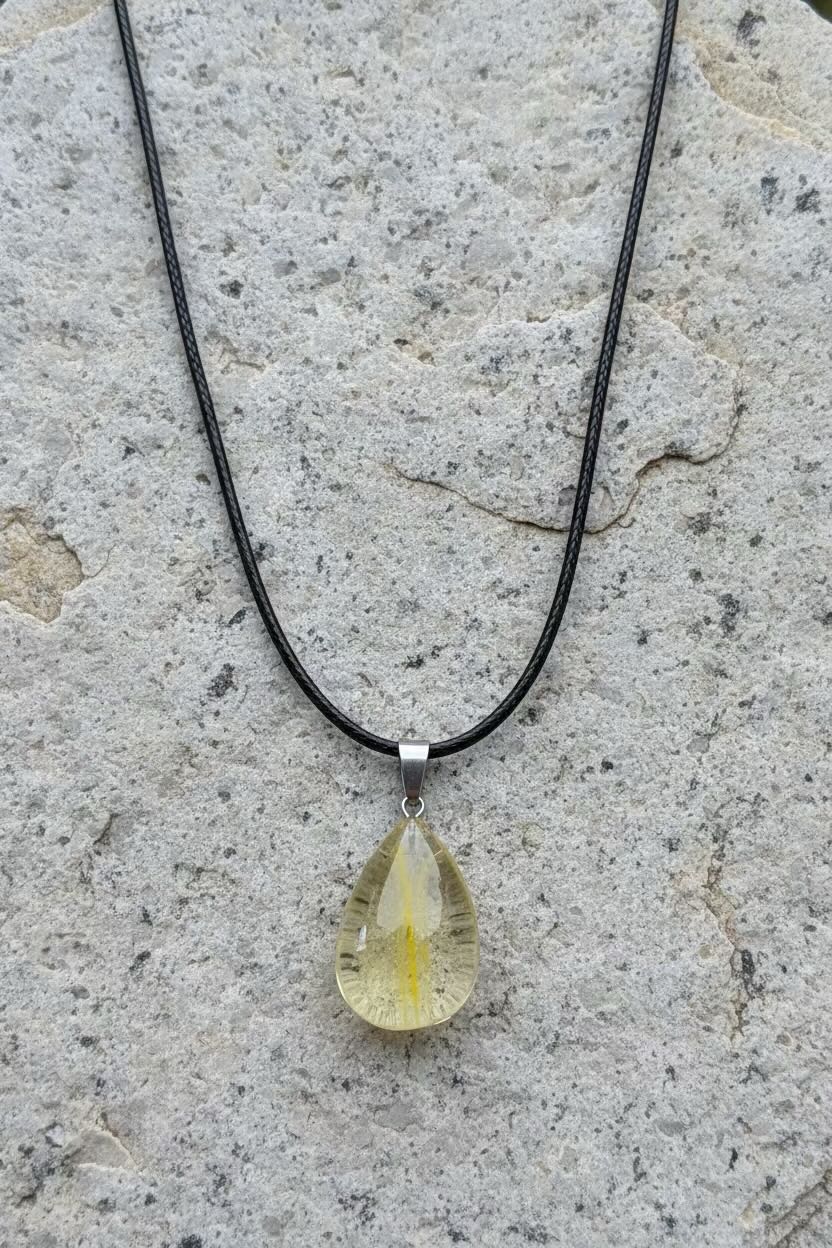 A teardrop-shaped yellow gemstone pendant on a black cord necklace, displayed on a textured gray stone surface.