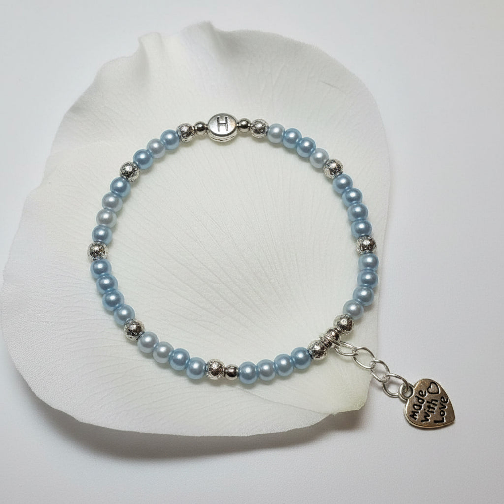 A beaded bracelet with light blue glass pearls and silver spacer beads, featuring a gold heart charm with a letter 'H' charm and a silver lobster clasp, shown on a plain light background.