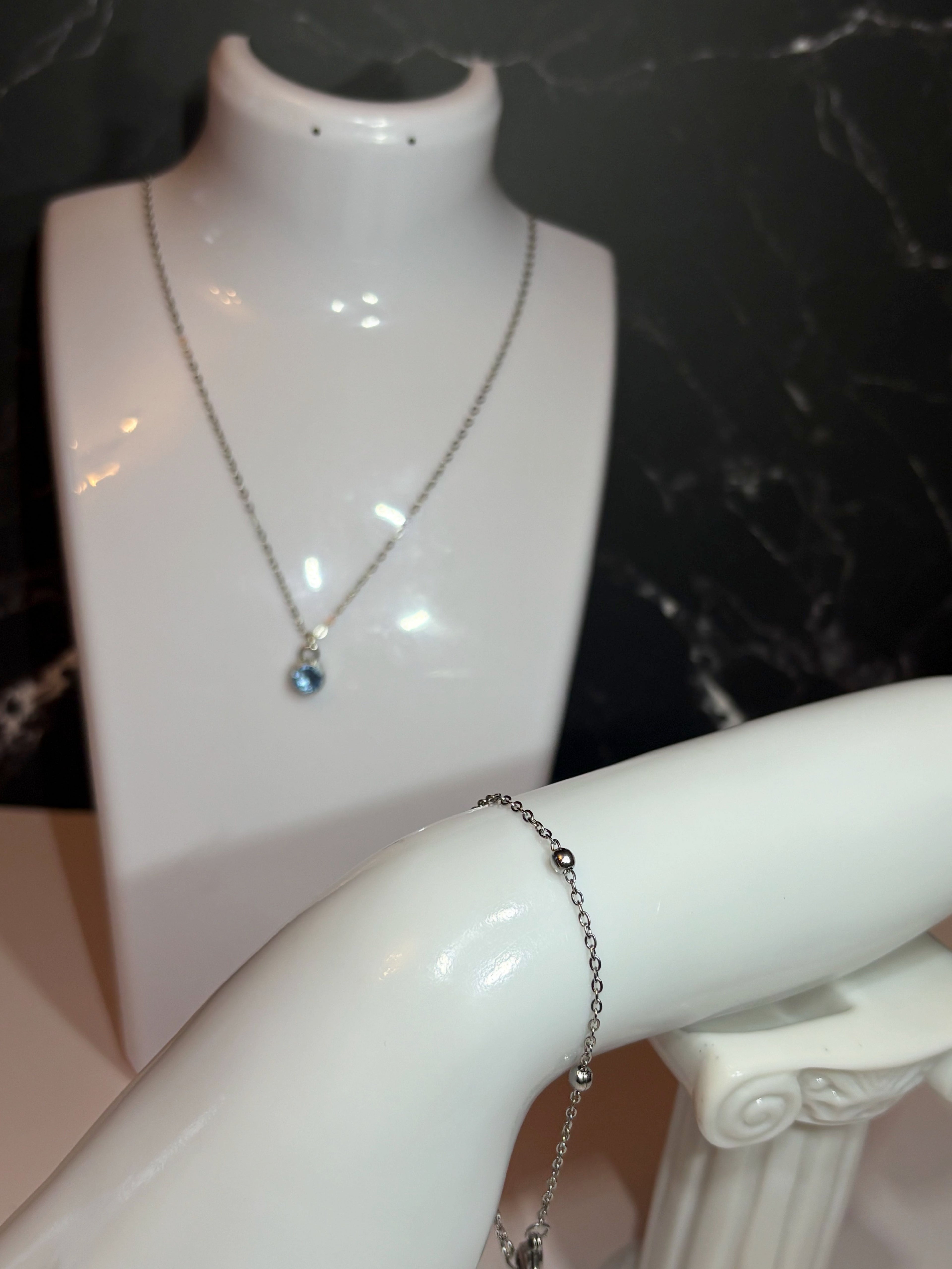 A silver-toned chain necklace with a small blue birthstone pendant displayed on a white jewelry bust, alongside a silver ball chain bracelet on the same bust arm.