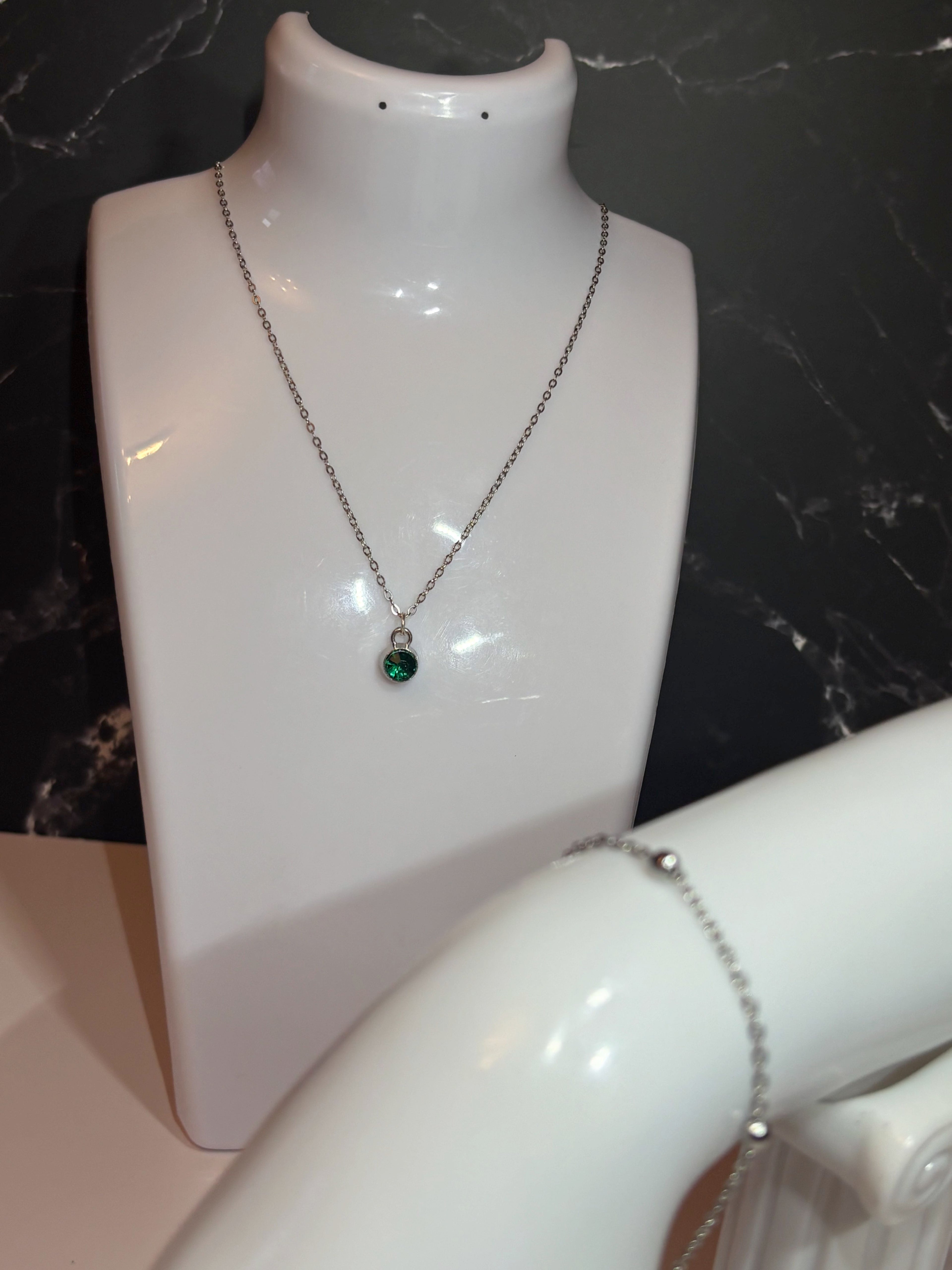 A silver chain necklace with a small round green birthstone pendant displayed on a white jewelry bust, with a silver ball chain bracelet partially visible on another arm stand against a dark marble background.