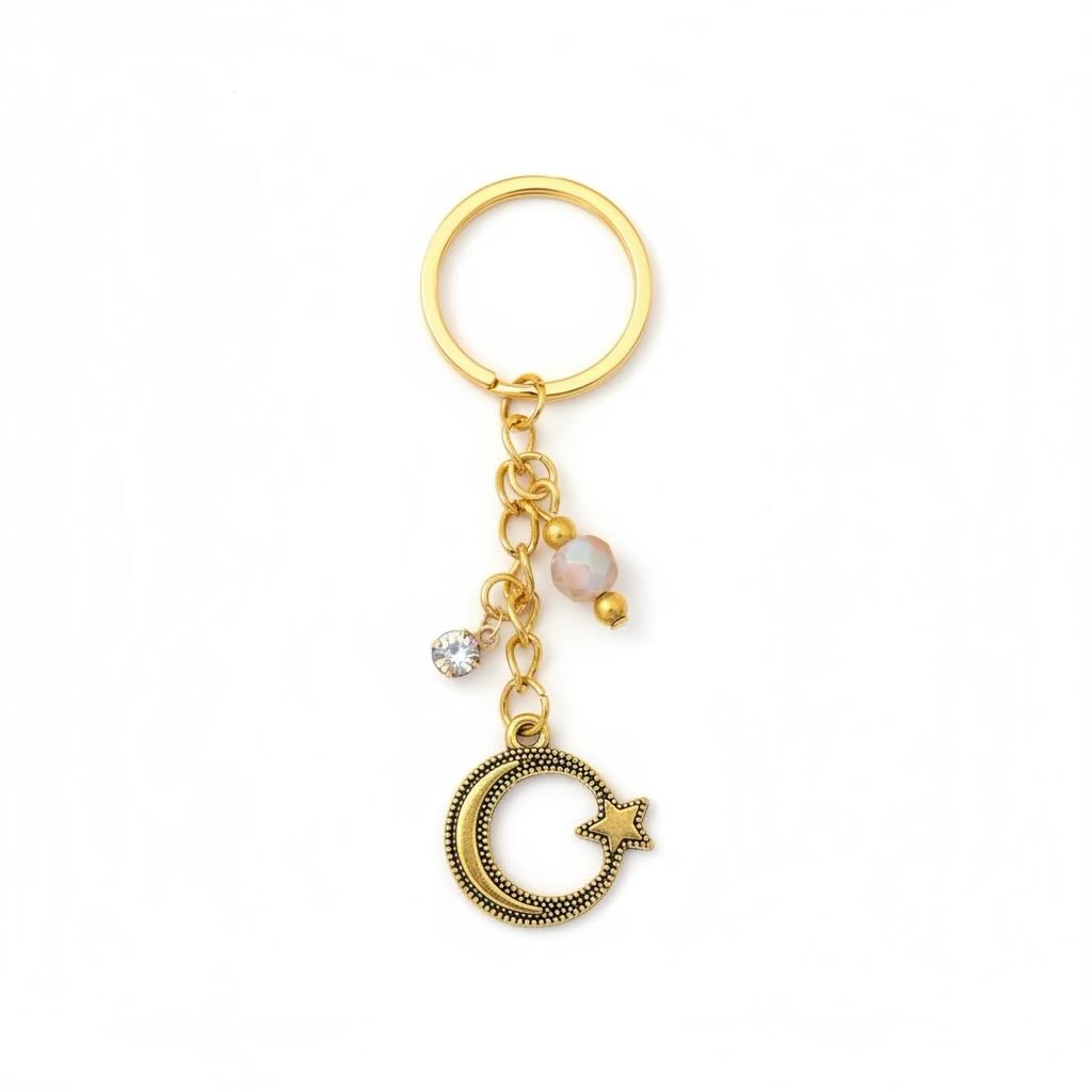 A gold-toned metal keyring featuring a round key ring, a short chain with a pale pink bead, a clear rhinestone charm, and a dangling crescent moon and star charm, shown on a plain white background.