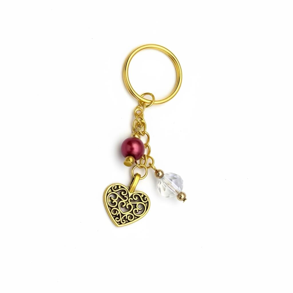A gold-toned filigree heart keyring with a dangling red bead and a clear faceted crystal charm, shown on a plain white background.