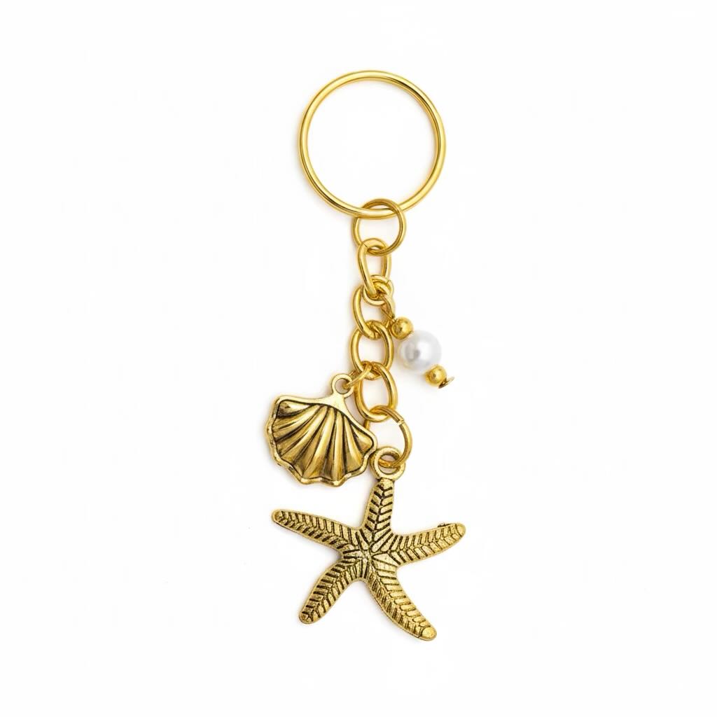 A gold-tone metal keyring featuring a starfish charm, a seashell charm, a small pearl bead, and a split key ring, shown on a plain white background.