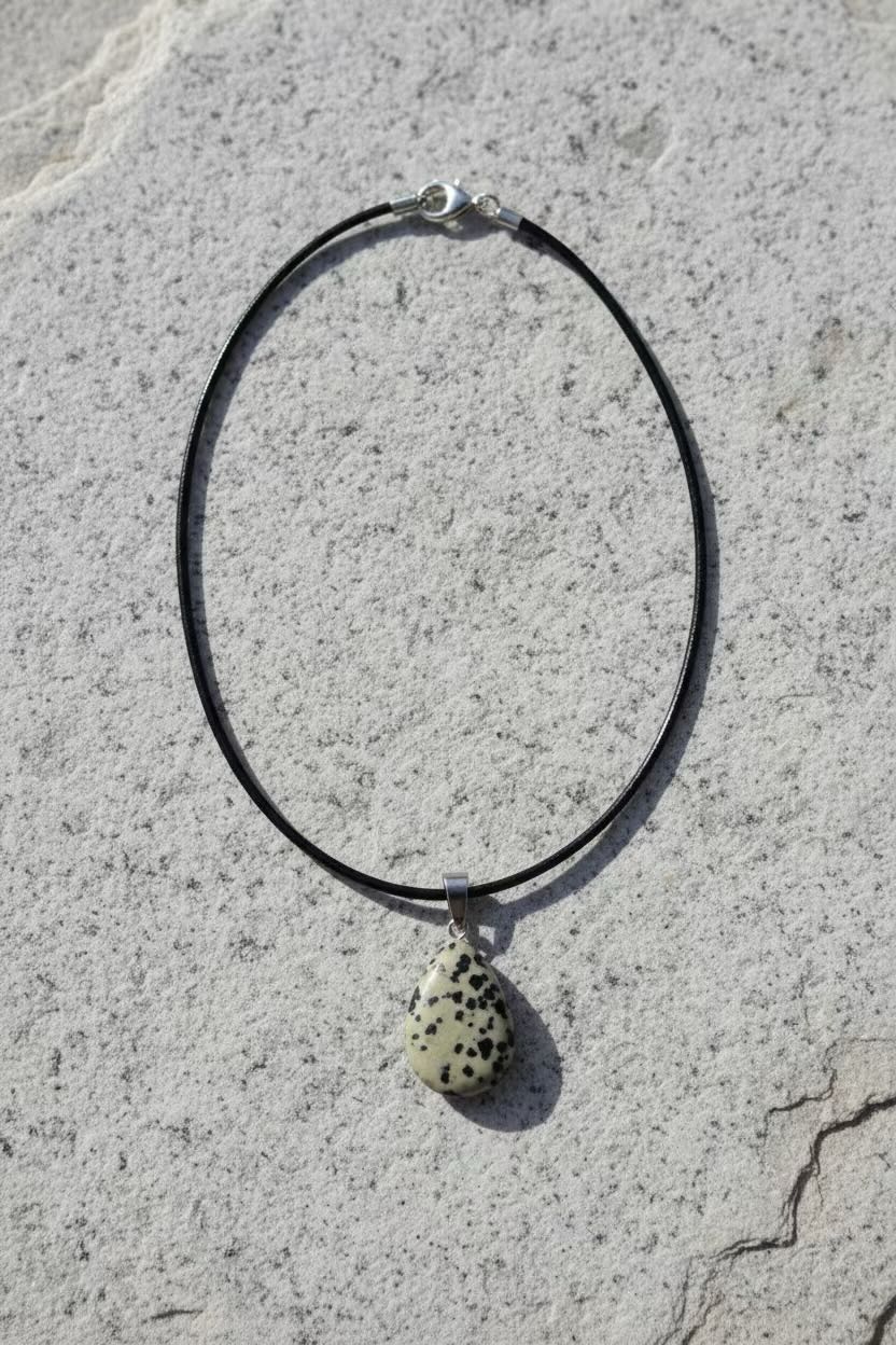 A black cord necklace with a small teardrop-shaped pendant featuring a black spotted pattern on a cream background, displayed on a textured gray stone surface.