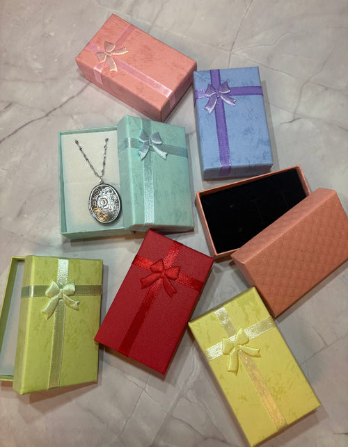A collection of colorful gift boxes and jewelry boxes in pink, blue, mint green, red, yellow, and orange, with one mint green box displaying a silver locket necklace on a chain, all arranged on a light marble surface.